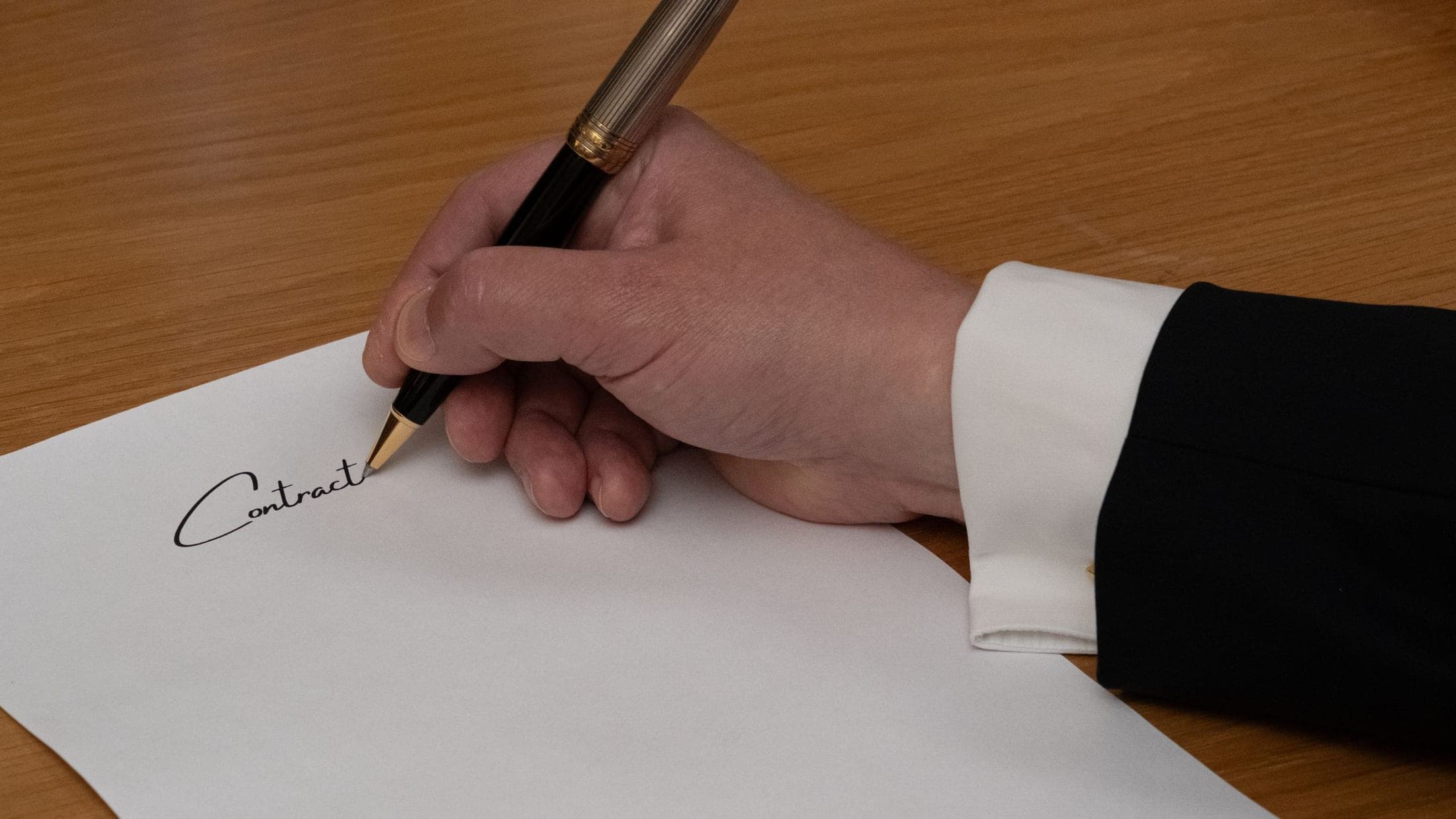 A hand signing a contract with a fountain pen at a wooden desk