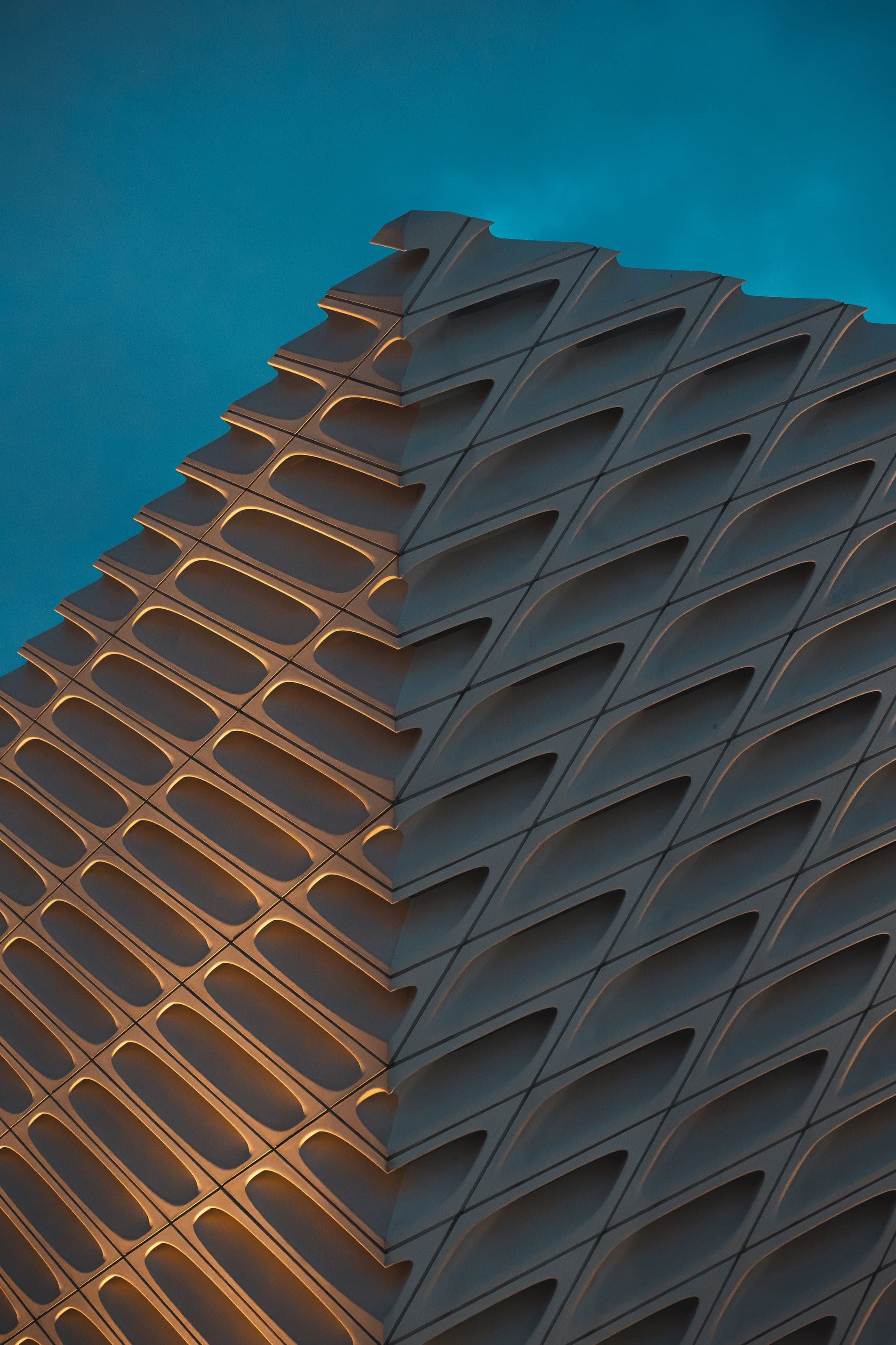 A geometric modern building facade with recessed panels lit by warm evening light against a teal sky